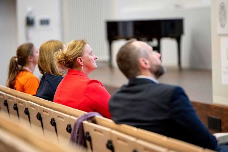 Mehrere Personen sitzen in einem Hörsaal auf hellen Holzbänken. Im Hintergrund steht ein schwarzer Flügel auf einer Bühne. Die Personen sind von hinten und leicht seitlich zu sehen, sie tragen unterschiedliche Kleidung, darunter ein auffällig rotes Jackett. Die Gesichter sind unscharf dargestellt.