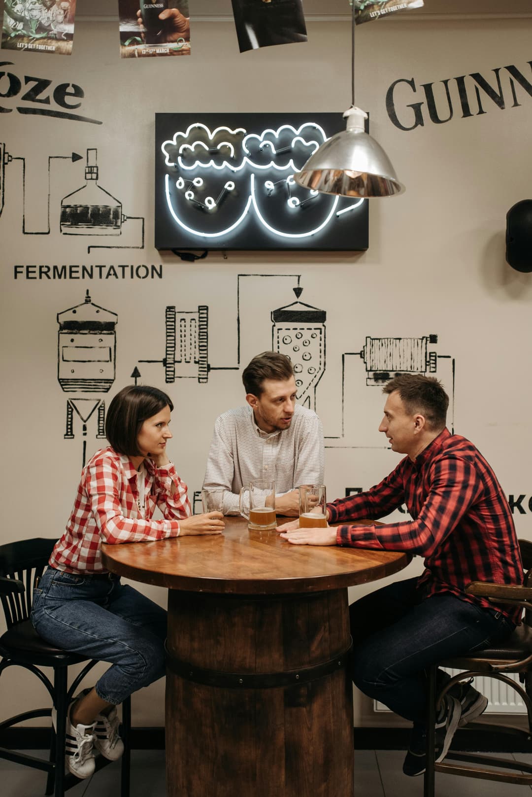 Drei Personen sitzen an einem runden Holztisch in einem Café oder einer Bar. Zwei tragen karierte Hemden, eine Person ein helles Hemd. Auf dem Tisch stehen Gläser mit Getränken. Die Wand im Hintergrund ist mit technischen Zeichnungen von Brauanlagen, englischen Begriffen wie 'FERMENTATION' und 'GUINNESS' sowie einer leuchtenden Wandinstallation dekoriert. Eine silberne Hängelampe beleuchtet die Szene.