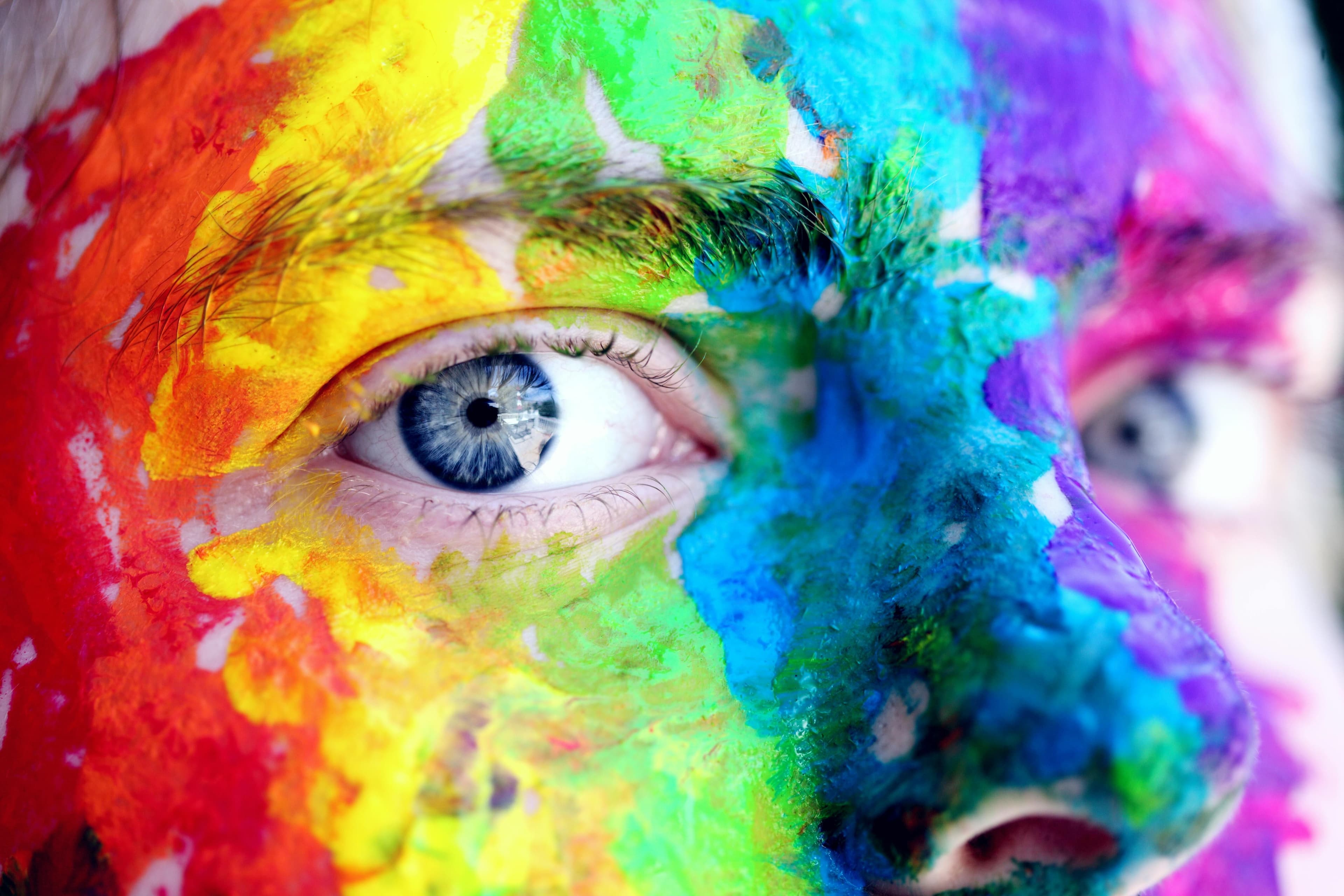 Nahaufnahme eines Gesichts mit intensiver, bunter Bemalung. Die Farben verlaufen in kräftigen Streifen und Flecken über die Haut und reichen von Gelb, Rot und Orange bis zu Blau, Grün und Violett. Das Auge in der Bildmitte ist klar erkennbar und wirkt durch die Bemalung besonders hervorgehoben. Die Struktur der Farbe ist stellenweise dick und unregelmäßig aufgetragen.