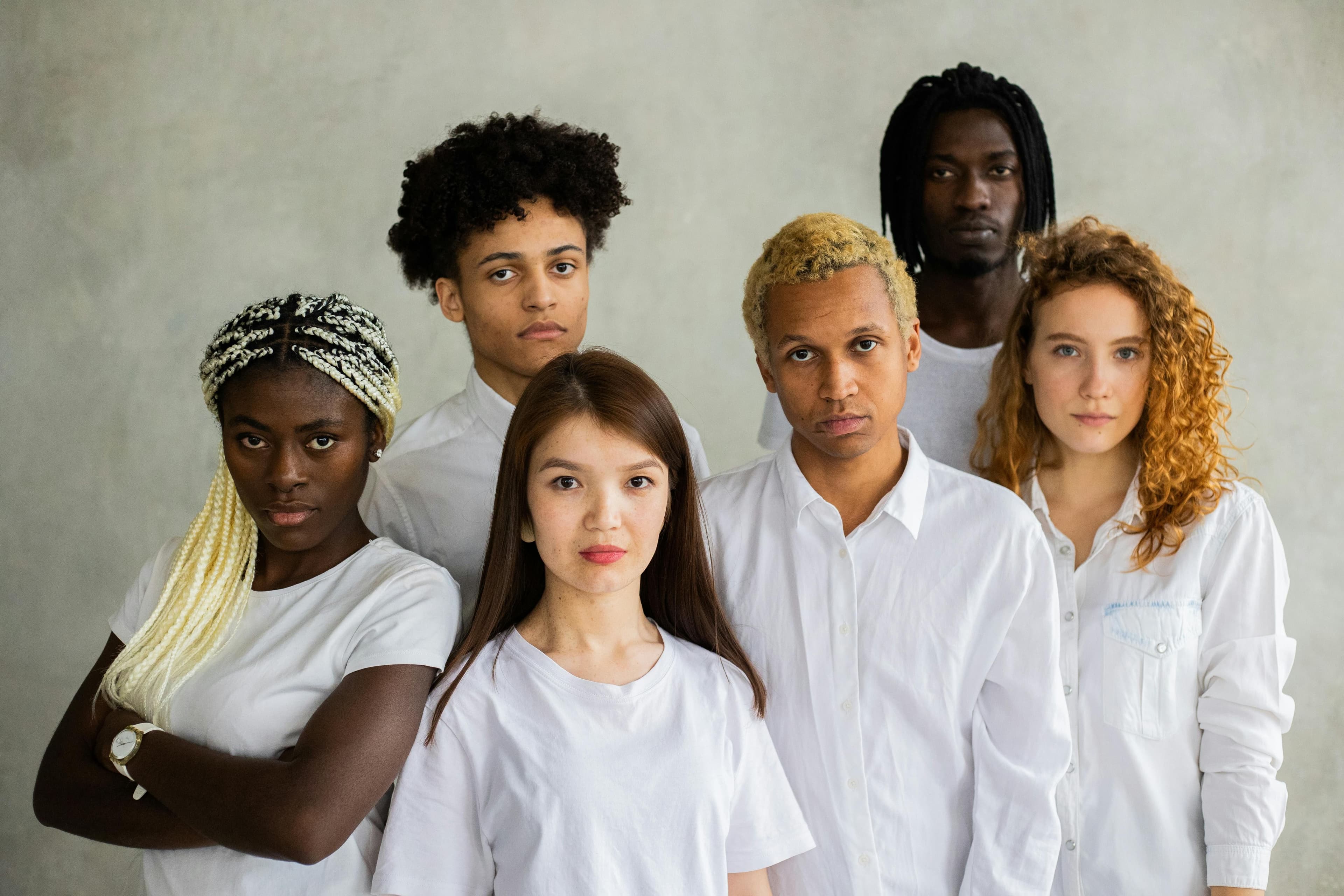 Sechs Personen stehen nebeneinander vor einem hellgrauen Hintergrund. Sie tragen weiße Oberteile und haben unterschiedliche Haarfarben und -strukturen. Die Körperhaltung variiert: Einige stehen mit verschränkten Armen, andere mit den Armen seitlich am Körper. Die Kleidung ist schlicht und einheitlich gehalten.