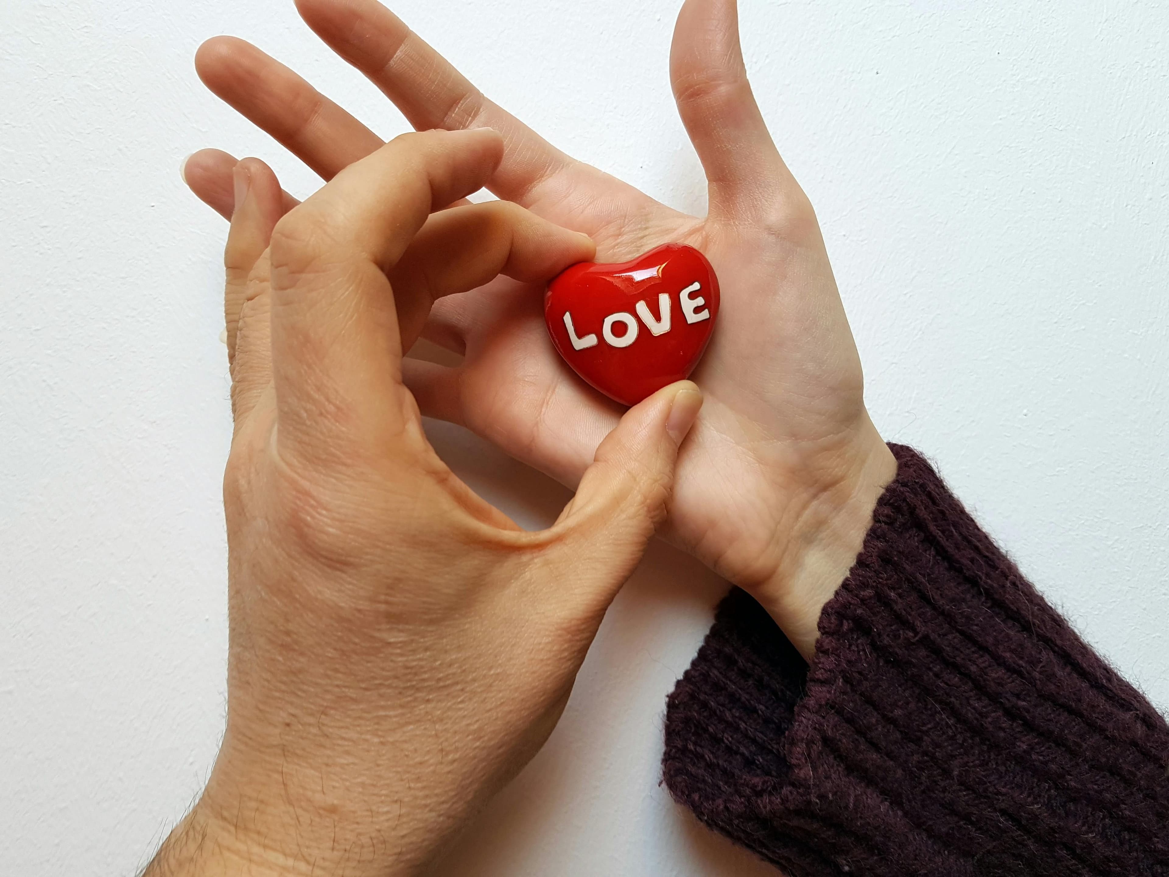 Zwei Hände halten gemeinsam ein rotes Herz mit der weißen Aufschrift 'LOVE'. Eine Hand trägt einen dunkelroten Strickpullover, die andere ist unbedeckt. Die Hände bilden eine schützende Geste um das Herz vor einem weißen Hintergrund.