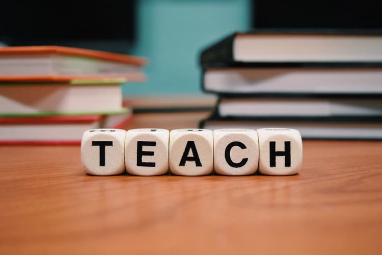Würfel mit den Buchstaben T E A C H auf einem Holztisch vor unscharfem Hintergrund mit mehreren Bücherstapeln